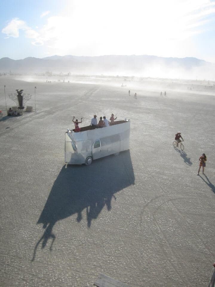 Aerial view — the mirror van nearly invisible on the playa, riders on top, dust haze